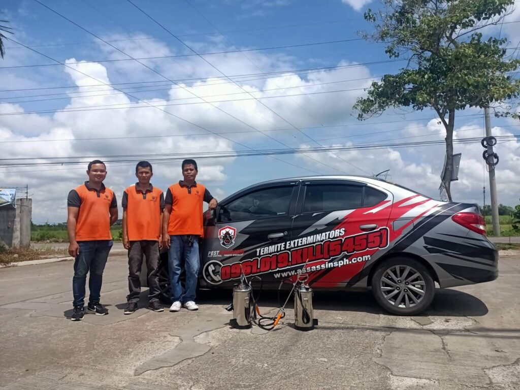 Three individuals in orange and black uniforms stand beside a black and red car branded with 'TERMITE EXTERMINATORS' and promotional details including a phone number and website. Two metal canisters, likely pest control equipment, are placed in front of the car. The setting is outdoors with trees, power lines, and a clear sky in the background.