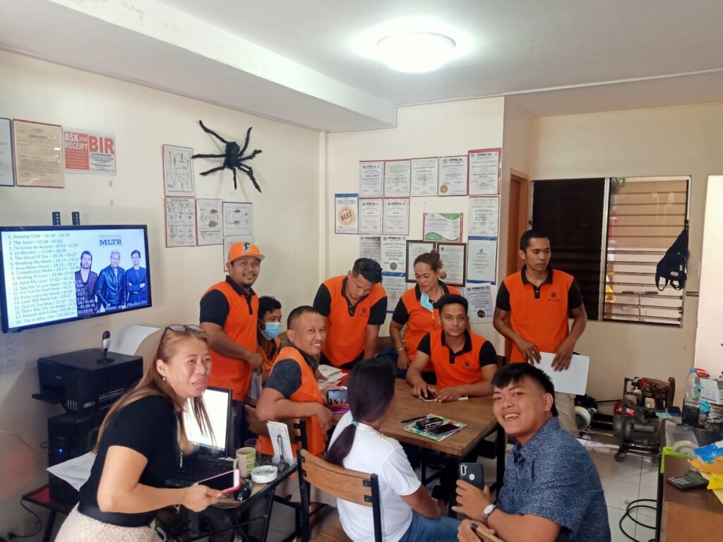 Team members of Pest Assassins Pest Control in orange uniforms attending a training session in a decorated office with certificates and a presentation screen.