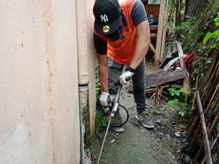 Pest control technician in orange vest and gloves uses pressure washer to clean a narrow outdoor passageway between a building wall and wooden fence, removing debris and vegetation.